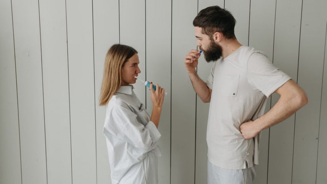 A couple brushing their teeth indoors, sharing a morning routine. Side view highlights interaction.