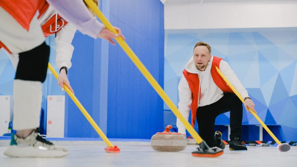 Determined sportsman throwing heavy granite curling stone near crop anonymous team sweeping ice sheet with brooms while playing sportive game