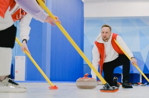 Determined sportsman throwing heavy granite curling stone near crop anonymous team sweeping ice sheet with brooms while playing sportive game