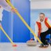 Determined sportsman throwing heavy granite curling stone near crop anonymous team sweeping ice sheet with brooms while playing sportive game